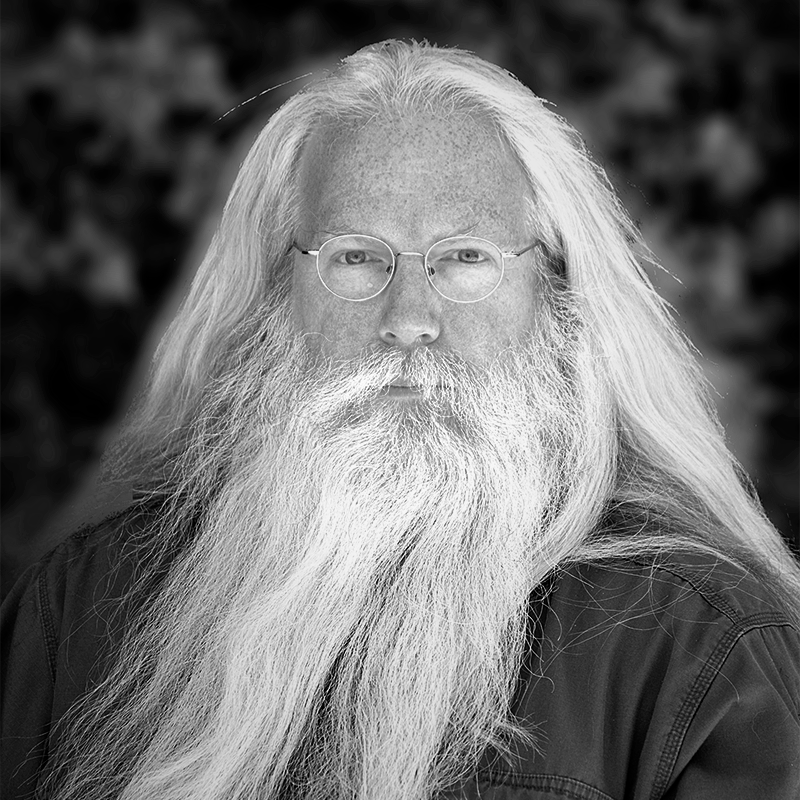 Michael is a light skinned man with long flowing white hair and a long beard, his face is freckled, and he is wearing round thin rimmed glasses and a dark shirt.